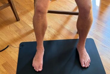 A person resting their bare feet on a black grounding mat placed on the floor, with the connection cord leading towards a wall outlet