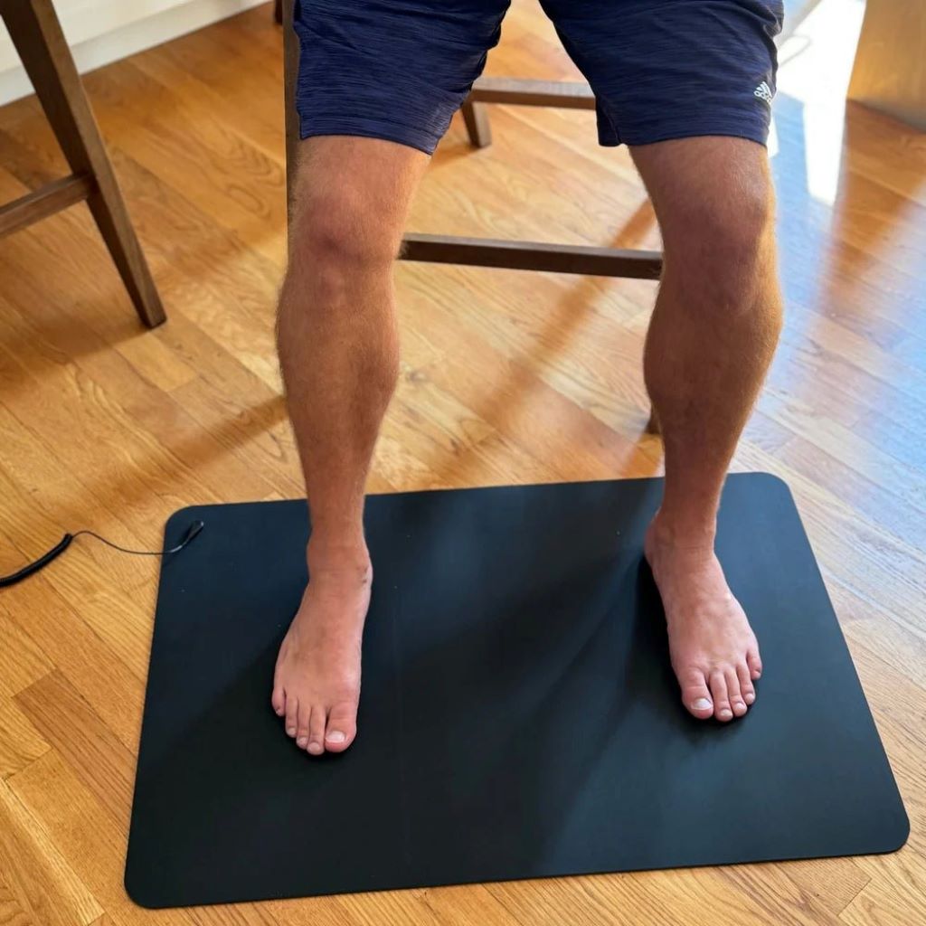 A person resting their bare feet on a black grounding mat placed on the floor, with the connection cord leading towards a wall outlet